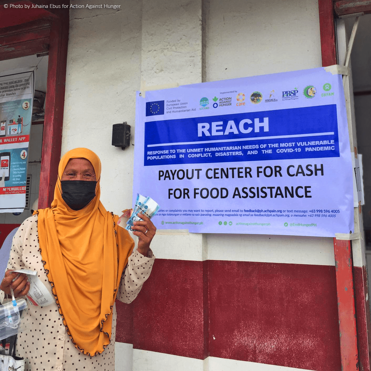 Cash Assistance Payout: Muslim woman holds cash paper bills while standing in front of REACH 2 project banner.