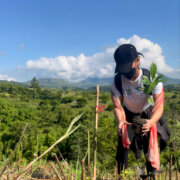 Female person in Action Against Hunger shirt wearing face mask and cap, holds a small tree to plant on the ground. Around her are green trees and bright blue sky.