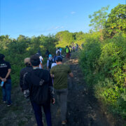 Individuals trekking a muddy dirt road, following a disorganized line.. Around them are green trees and bright blue sky.