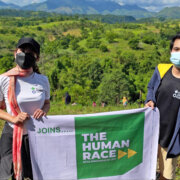 Two individuals carrying a white banner with the text "Joins..." along with #TheHumanRace logo (Green square with bold white capitalized text 'THE HUMAN RACE' followed by two yellow arrows, and a small text a the bottom 'World Humanitarian Day 2021'
