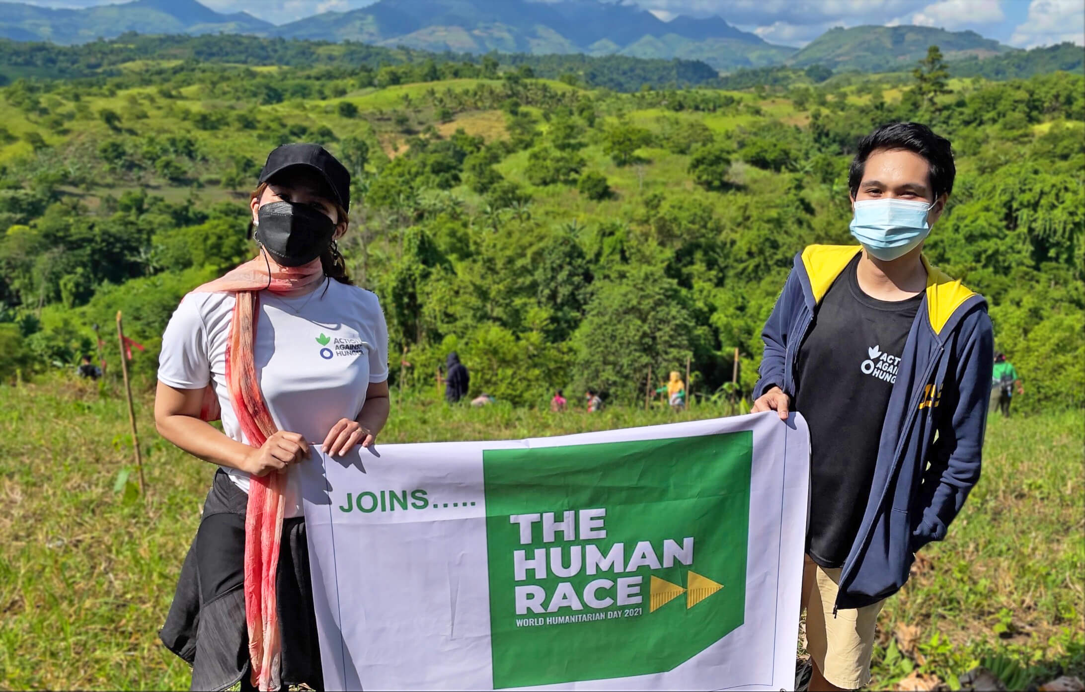 Two individuals carrying a white banner with the text "Joins..." along with #TheHumanRace logo (Green square with bold white capitalized text 'THE HUMAN RACE' followed by two yellow arrows, and a small text a the bottom 'World Humanitarian Day 2021'
