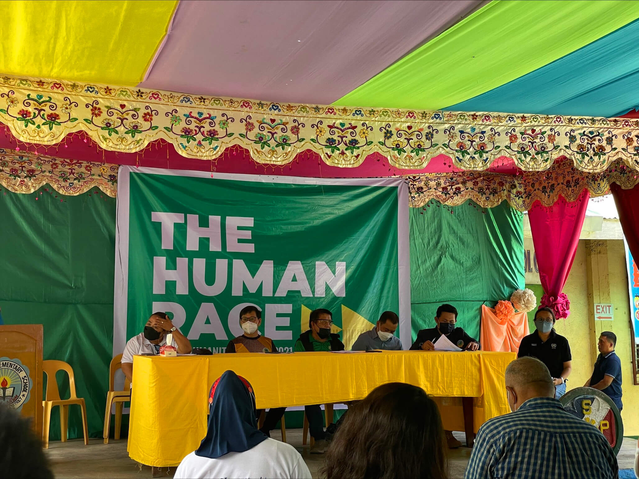Six individuals sitting in a long table covered in yellow table cloth. Behind them is a green backdrop tarpaulin with bold text " THE HUMAN RACE" An audience is seated in front of them.