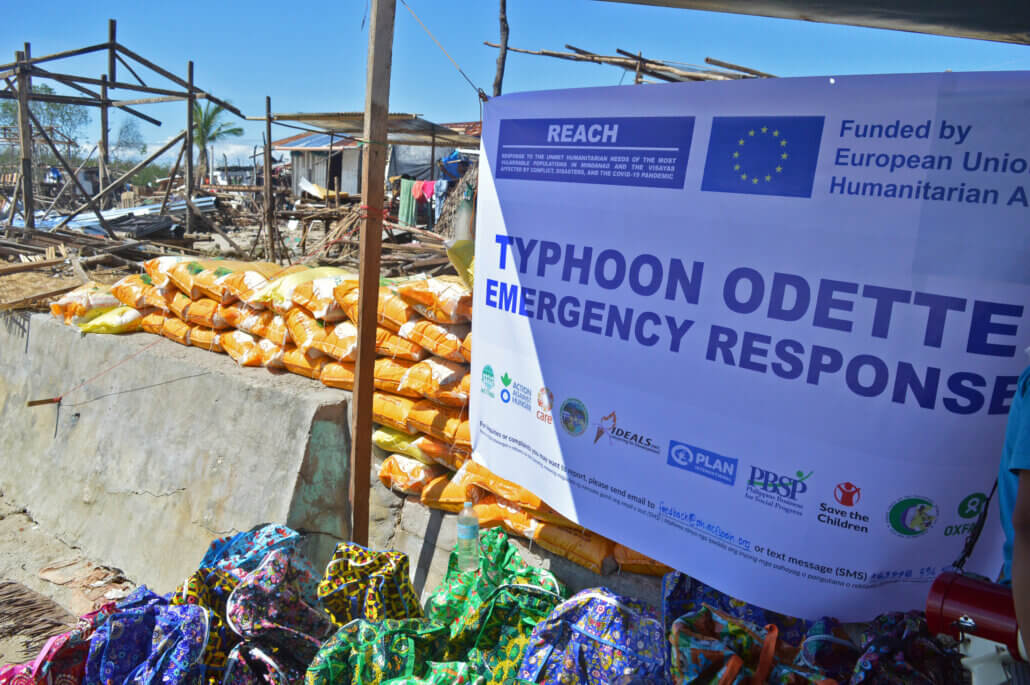 Food packs and NFI kits are lined up along the entrance of the barangay; a REACH Banner hangs and behind it are broken houses.