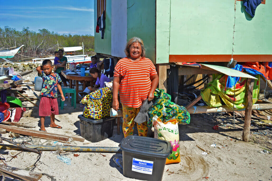 Flora Cuaton smiles upon receiving the food packs and NFI kits from REACH - Typhoon Odette Emergency Response. On the left side of the photo are the debris from their damaged house. The green house behind her is owned by one of their children, where they now temporarily stay altogether.