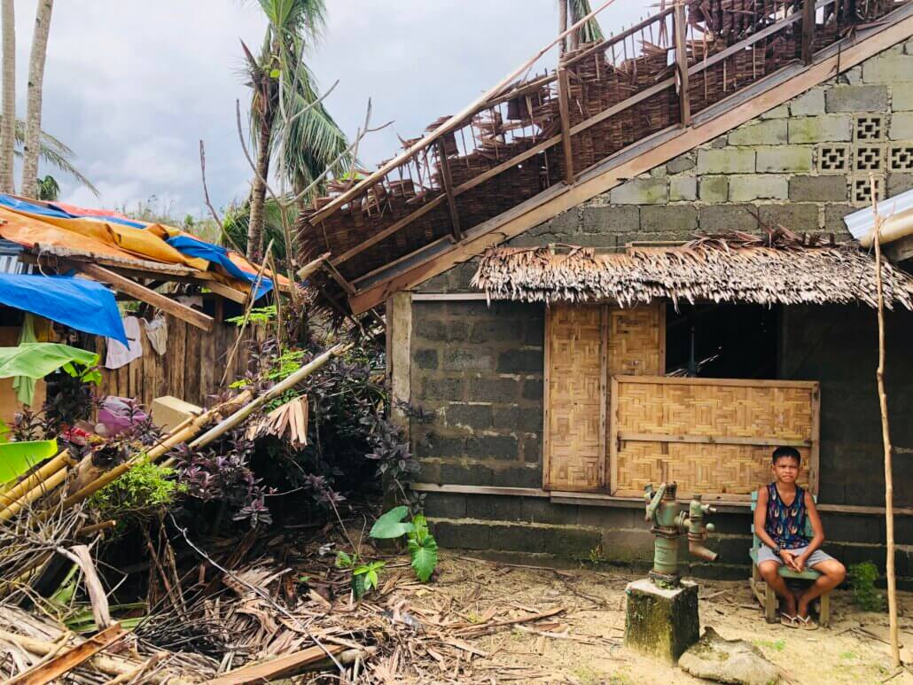 Boy sitting outside his house; house is a combination of concrete, wood and bamboo. The roof is partially damaged